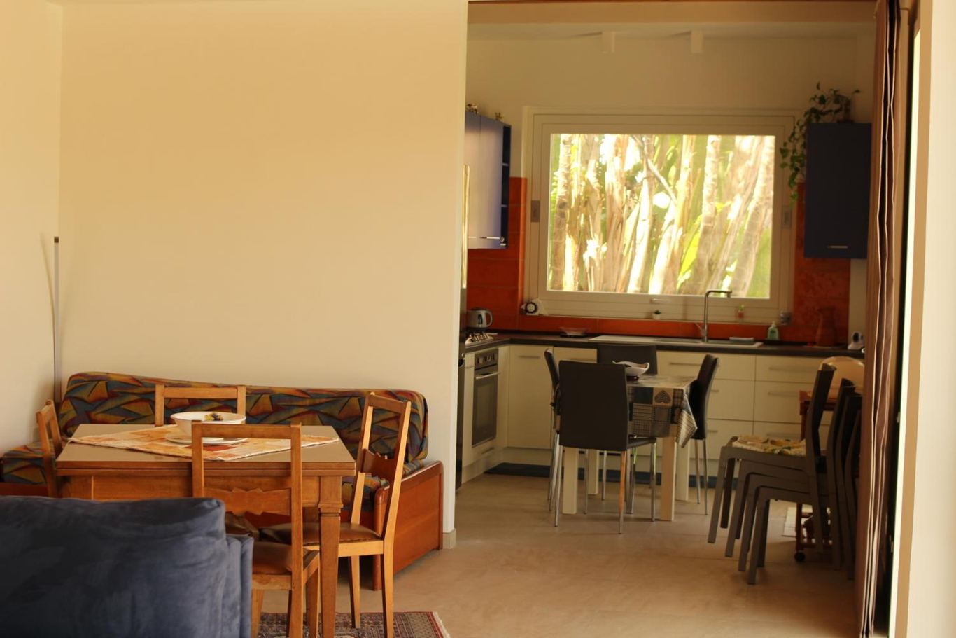 Il living con il divano letto e il tavolo, e la cucina in fondo con la finestra sulle palme.