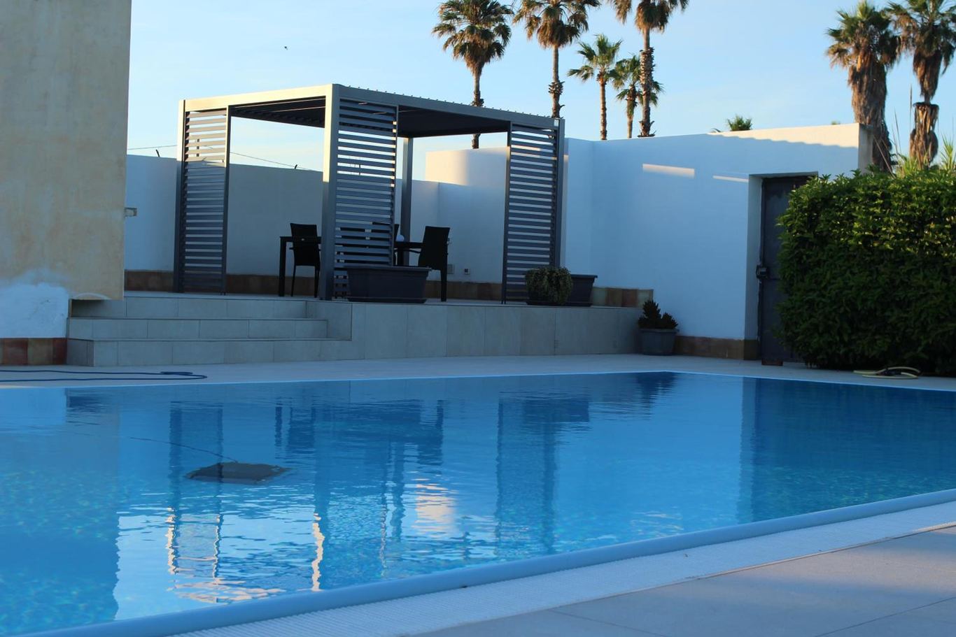 La pergola e la piscina nel sole del pomeriggio, con le palme dietro il muro.