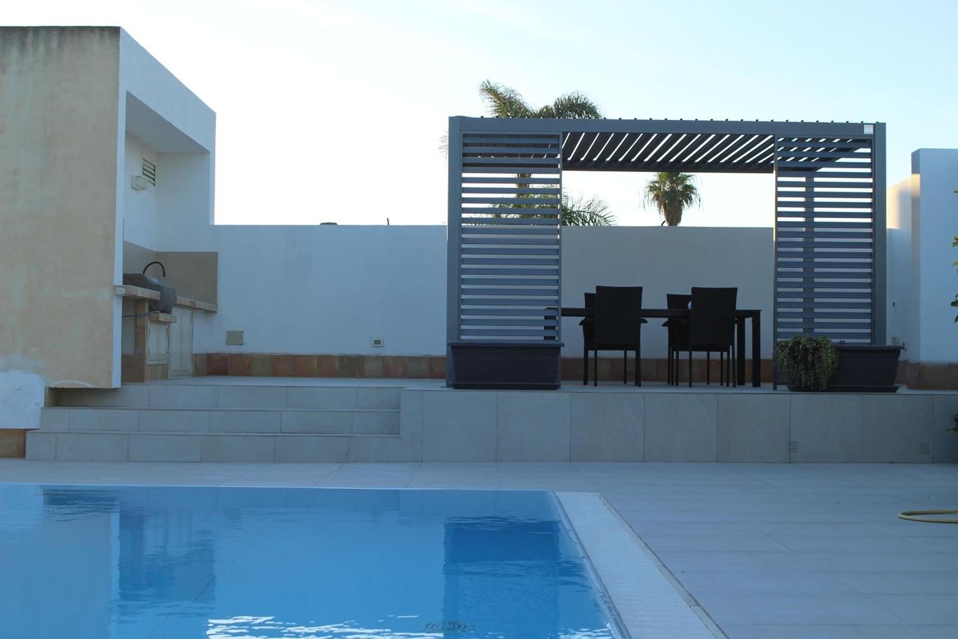 La piscina con la pergola metallica e il tavolo da pranzo sul solarium.
