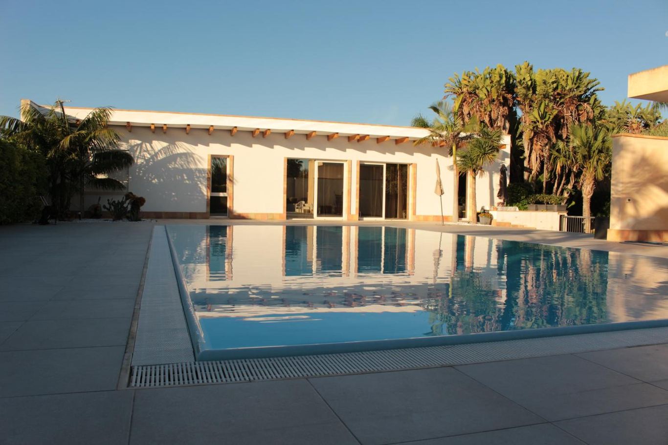 La piscina a sfioro con la villetta bianca e le palme sullo sfondo, nella luce calda del tramonto.