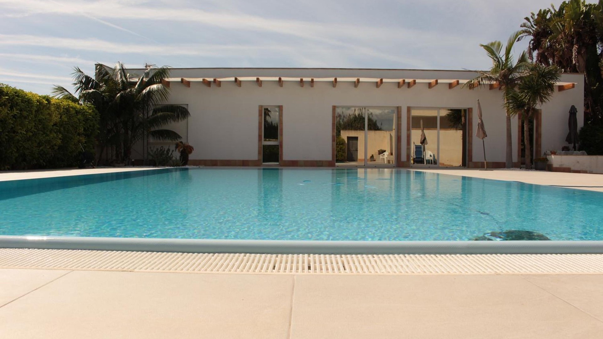 Villetta Nonna Giovanna a Marsala: la casa bianca con la pergola di legno, la piscina a sfioro, il solarium, le palme e il giardino.